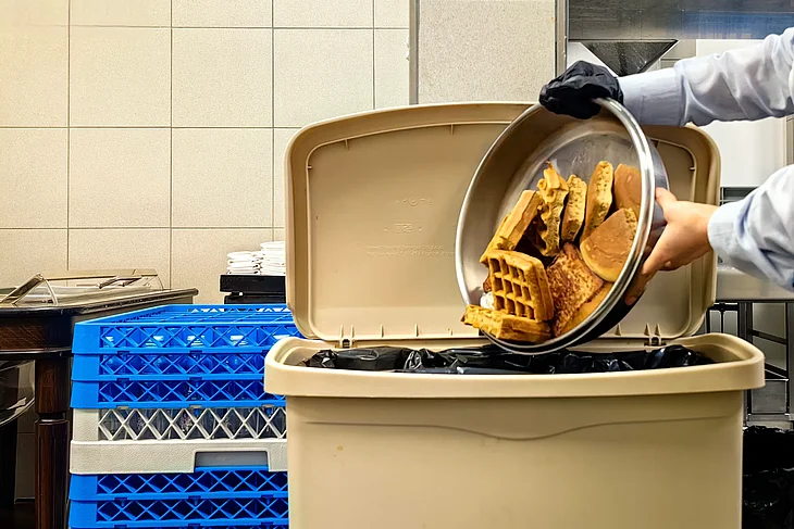 Ein Teller mit Gebäck wird in den Bioabfall gekippt – Symbolbild für Lebensmittelabfälle, die in dem Artikel als Ausgangsstoff für die Gewinnung von Rohstoffen mithilfe von Sonnenlicht thematisiert werden.